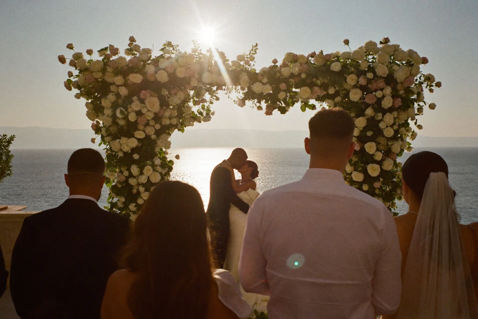 Primo bacio sotto un arco di rose di fronte al mare, visto da dietro gli ospiti
