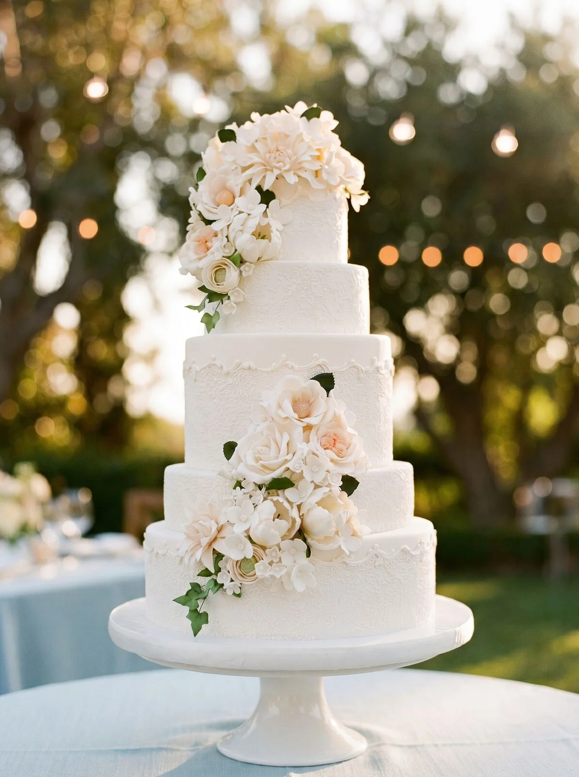 Torta nuziale bianca a piani, cascata di fiori crema e cipria, giardino di sera